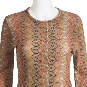 Puede incluir: Un cárdigan con botones en una cálida paleta de colores marrón, naranja y amarillo. El tejido de punto presenta un patrón repetitivo en forma de hoja y un cuello redondo. El suéter de manga larga tiene cinco botones a juego en la parte delantera.