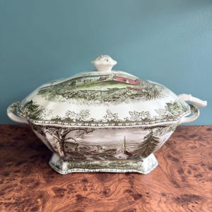 Pode incluir: Uma terrina de cerâmica cor de creme com tampa, apresentando uma estampa detalhada de paisagem em tons de verde e castanho. O design retrata uma cena rural com um celeiro vermelho, uma igreja, árvores e colinas, com bordas florais decorativas.