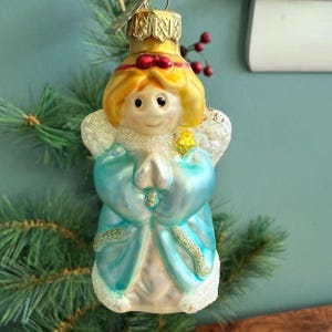 May include: A vintage Christmas ornament shaped like an angel. The angel has blonde hair, a red bow, and a light blue robe. The ornament has glitter accents and is holding its hands in prayer. A gold-colored metal cap tops the ornament.