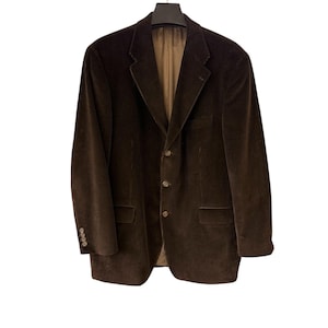 Puede incluir: Americana de pana marrón oscuro con cierre de tres botones y dos bolsillos delanteros. La americana está colgada de una percha negra sobre un fondo blanco. Tiene solapas con muescas y puños abotonados.