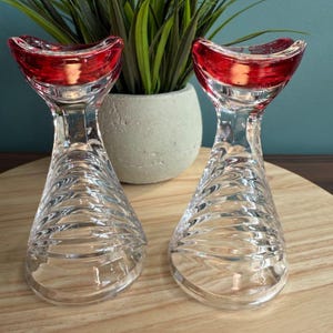Puede incluir: Dos candelabros de cristal transparente con una parte superior roja y curvada. La base de cada soporte tiene un diseño texturizado y arremolinado. Los candelabros están sobre una superficie de madera, con una planta en maceta en el fondo.