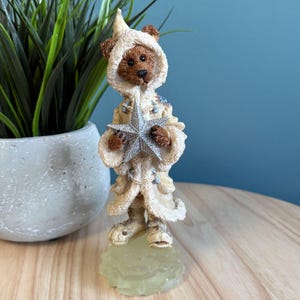 Pode incluir: Uma estatueta de um urso marrom vestindo um casaco com capuz e botas creme, segurando uma estrela prateada. O urso está em uma base verde clara. O casaco tem acabamento em pele sintética e detalhes azuis. A estatueta está em uma superfície de madeira.