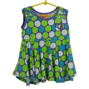 May include: Sleeveless dress with a purple, green, teal, and white polka dot pattern. The dress has a flared skirt and a purple neckline. The shoulders are decorated with green and white fringe. A sequined patch is on the chest. The dress is hanging on a yellow hanger.