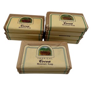 May include: Several bars of Tropical Cocoa Moisture Soap are stacked and arranged on a white surface. The soap is wrapped in brown paper with a green and brown label featuring a tropical scene. The text "Tropical Cocoa Moisture Soap" is printed on the label.