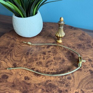 May include: A gold-toned lamp harp and finial sit on a wooden surface. The harp is a curved metal piece with a screw and two pointed ends. The finial is a decorative, gold-colored piece with a rounded top and a fluted design.