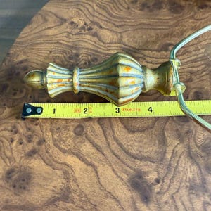 May include: A gold-colored, ornate finial with a decorative design, measuring approximately 4 inches in length. The finial has a distressed finish and is placed on a wooden surface. A yellow measuring tape is next to the finial.
