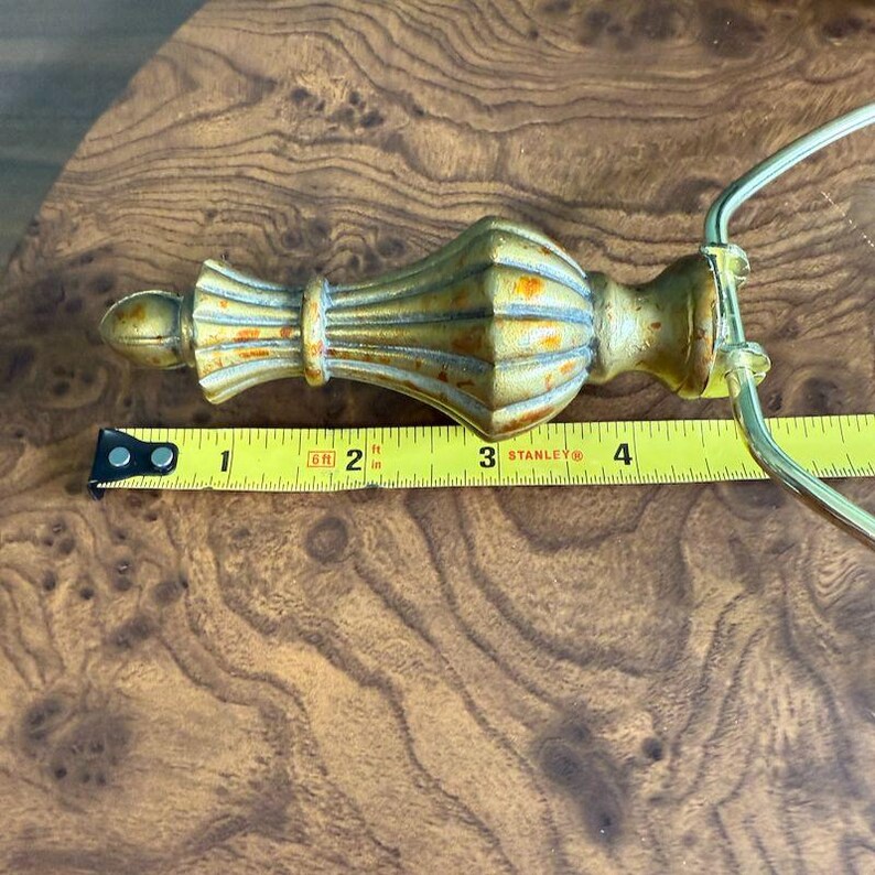 May include: A gold-colored, ornate finial with a decorative design, measuring approximately 4 inches in length. The finial has a distressed finish and is placed on a wooden surface. A yellow measuring tape is next to the finial.