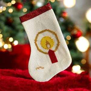 Puede incluir: Calcetín navideño color crema con un diseño cosido de una vela amarilla dentro de un círculo dorado y una base roja. La parte superior del calcetín tiene un ribete rojo estampado. El año "1996" está cosido en dorado en la parte inferior.