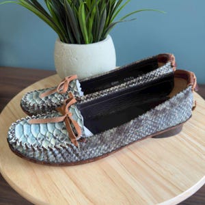 Peut inclure: Une paire de mocassins à imprimé serpent avec des écailles bleu clair et grises, des bordures en cuir marron et de petits nœuds. Les chaussures ont un bout arrondi et un petit talon. Les mocassins sont présentés sur une surface en bois.