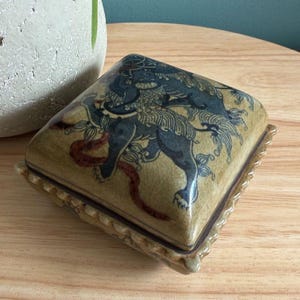 May include: A square, ceramic trinket box with a detailed illustration of a mythical creature in shades of blue and tan. The box has a decorative, ridged border and a matching lid. The design is reminiscent of Asian art.