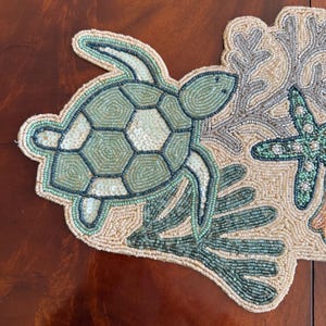 May include: A beaded placemat with a sea turtle, starfish, and coral reef design. The turtle has a green and white shell, and the starfish is green. The placemat is beige and has a dark brown border.