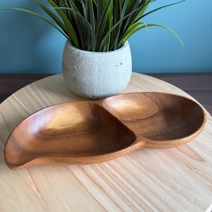 Puede incluir: Una fuente de madera con dos compartimentos conectados. La fuente es de madera marrón claro y tiene un acabado liso y pulido. Una pequeña maceta gris con una planta verde se encuentra en el fondo.