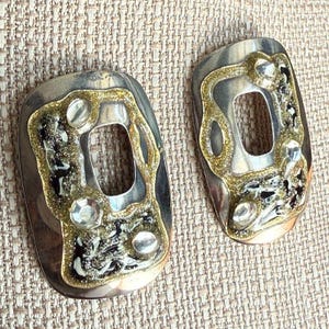 May include: A pair of silver-toned earrings with an abstract design. Each earring features a rectangular shape with a central cutout, accented with gold-colored trim, clear stones, and black and silver detailing.