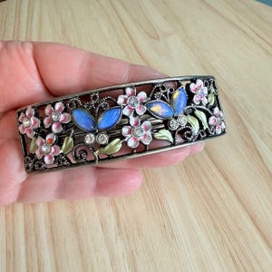 Könnte beinhalten: Eine rechteckige Haarspange mit einem dunklen Metall-Filigranrahmen, verziert mit rosa Emailleblumen, grünen Emailleblättern und zwei blauen, schillernden schmetterlingsförmigen Cabochons. Das Design enthält kleine klare Strasssteine in jeder Blütenmitte.