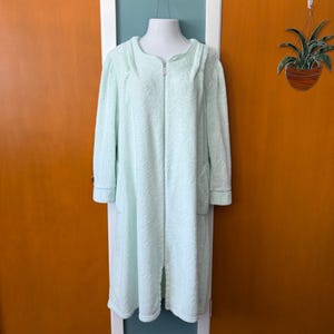May include: A light green, full-length robe with a hood and zipper closure. The robe has long sleeves, pockets, and a subtle floral pattern. The robe is displayed on a mannequin and appears to be made of a soft, plush material.
