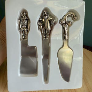 Peut inclure: Un ensemble de trois couteaux à fromage argentés. Chaque couteau présente un manche décoratif : un casse-noisette, le Père Noël et une canne en bonbon avec un nœud. Les couteaux sont présentés dans un plateau en plastique blanc.