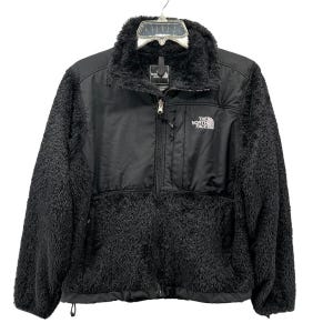 May include: Black fleece jacket with a zip-up front and a high collar. The jacket has a smooth, black panel across the chest and shoulders, with the brand logo on the upper right. The jacket is hanging on a hanger.
