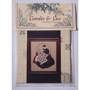 May include: A cross-stitch kit titled "Lavender & Lace Victorian Designs." The kit features a framed image of two figures, one in a dark dress and the other in a red dress, working on a patterned textile. The kit is packaged in a clear plastic sleeve.