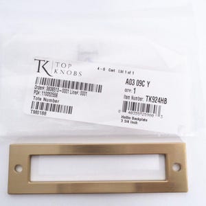 May include: A rectangular brass-toned Hollin backplate with a white insert and two screw holes. The backplate measures 3 3/4 inches. A label with the text "TOP KNOBS" and item details is in the background.