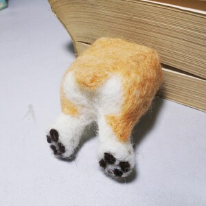 May include: A felt sculpture of a dog's rear end with white fur and brown spots on the paws. The dog is partially hidden behind a book.