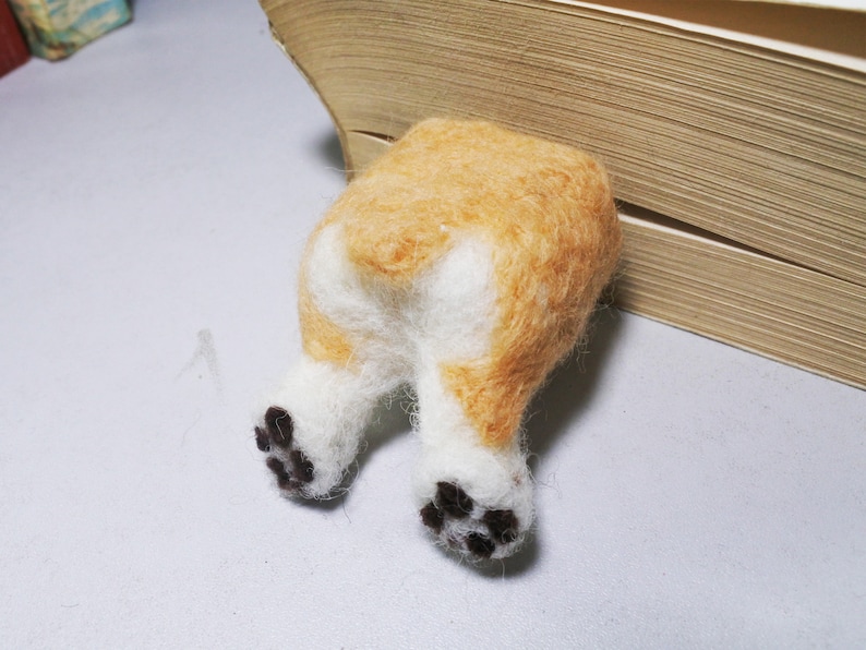May include: A felt sculpture of a dog's rear end with white fur and brown spots on the paws. The dog is partially hidden behind a book.