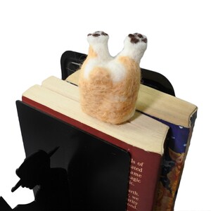 May include: A black metal bookend featuring a silhouette of a person reading. A small, felted animal resembling a dog is perched on top of the bookend, with its back legs in the air.