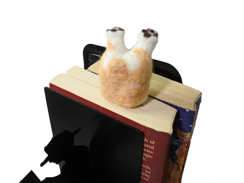 May include: A black metal bookend featuring a silhouette of a person reading. A small, felted animal resembling a dog is perched on top of the bookend, with its back legs in the air.