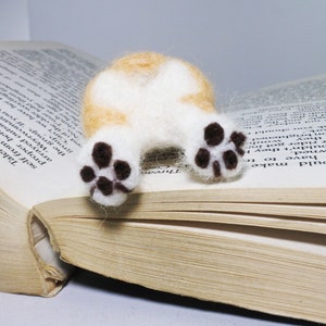 May include: A small, white and orange felt animal with brown paw prints, lying on an open book. The animal is a dog, and its tail is curled up.