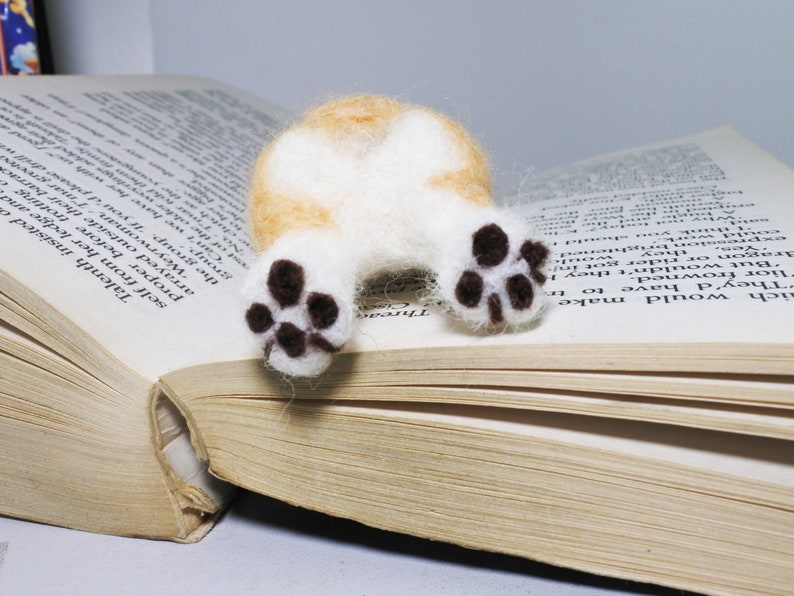 May include: A small, white and orange felt animal with brown paw prints, lying on an open book. The animal is a dog, and its tail is curled up.
