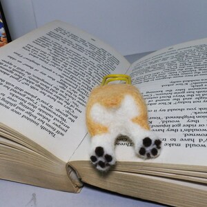 May include: A felt bookmark shaped like a corgi dog with brown and white fur. The bookmark is lying on a book with the text "Threac" and "Cise" visible.