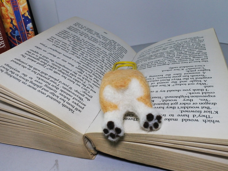 May include: A felt bookmark shaped like a corgi dog with brown and white fur. The bookmark is lying on a book with the text "Threac" and "Cise" visible.
