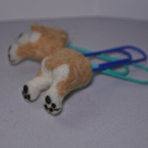May include: A small, brown and white felt dog figurine attached to two blue and green paper clips. The dog has black eyes and a black nose.