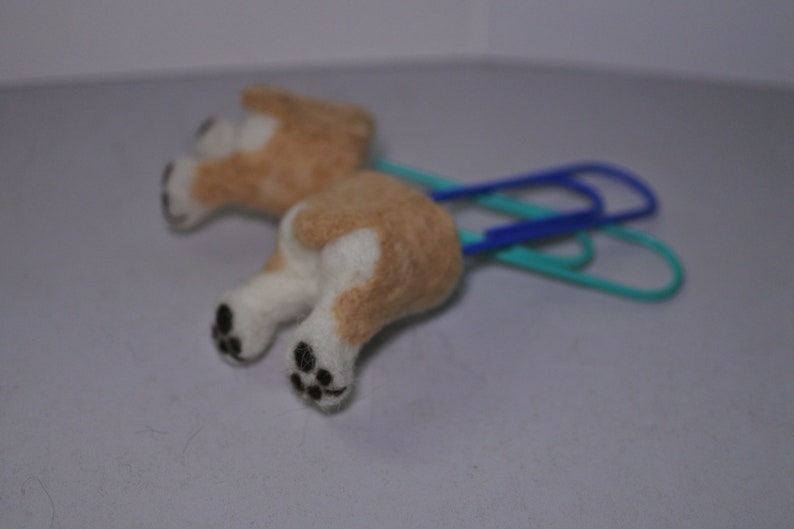May include: A small, brown and white felt dog figurine attached to two blue and green paper clips. The dog has black eyes and a black nose.