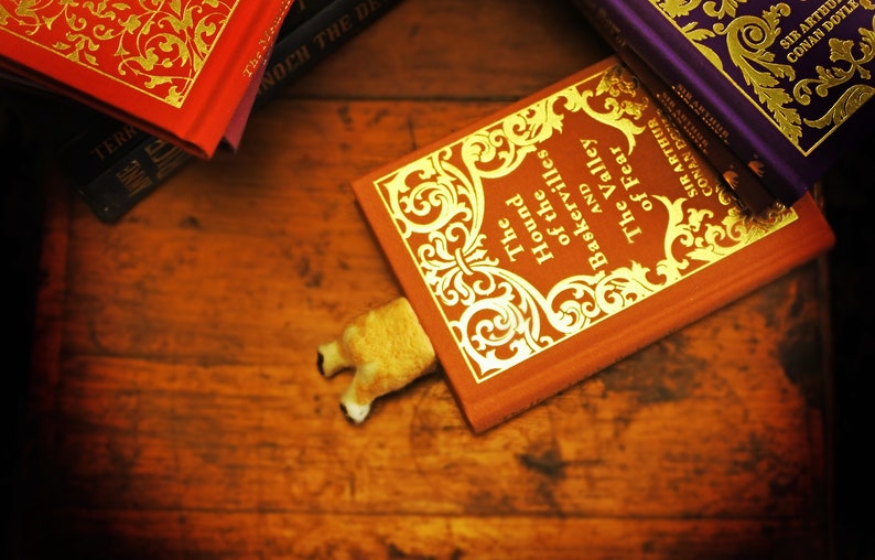 May include: A red hardback book with gold lettering titled "The Hound of the Baskervilles and The Valley of Fear" by Sir Arthur Conan Doyle. A small, brown, plush dog bookmark is sticking out from the book.