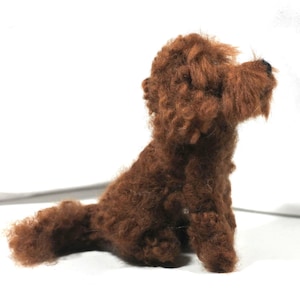 May include: A brown, fluffy, needle-felted dog toy. The dog is sitting on a white surface and is looking to the right.