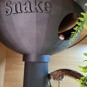 May include: A black plastic snake hide with the word "Snake" printed on the front. The hide has a round opening for the snake to enter.