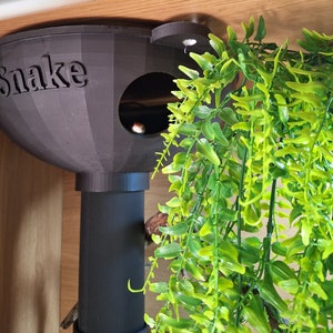 May include: A black plastic snake hide with the word "snake" printed on the front. The hide has a round opening and a tube extending from the bottom. A green artificial plant is hanging in the background.