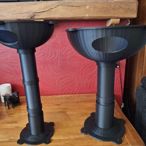 May include: Two black cat perches with a circular opening. The perches have a bowl-shaped top, a cylindrical support, and a round base. They are placed on a wooden surface against a red patterned wall.