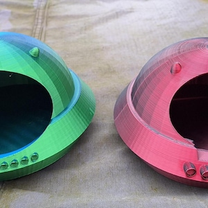 以下が含まれることがあります： 前面に円形の開口部がある、3Dプリントされたドーム型のオブジェクトが2つあります。1つは緑と青、もう1つは赤です。どちらも下端に小さな円形の突起が並んでいます。