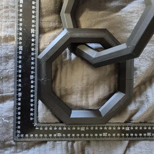 Op de afbeelding: Donkergrijze geometrische schakels, mogelijk 3D-geprint, zijn in elkaar verweven. Een zwarte liniaal met witte markeringen in centimeters en millimeters is naast de schakels geplaatst, voor de schaal. De schakels liggen op een grijze stoffen ondergrond.