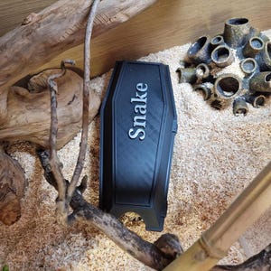Op de afbeelding: Zwarte plastic slangschuilplaats met het woord "Snake" in reliëf in witte letters.