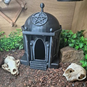 May include: A dark grey, gothic-style terrarium hideout with a pentagram on top. The hideout has arched doorways and is surrounded by faux greenery, soil, and animal skulls. The design is reminiscent of a small temple or mausoleum.