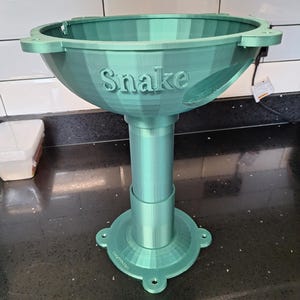 Peut inclure: Un support imprimé en 3D de couleur sarcelle avec le mot "Snake" sur le dessus en forme de bol. Le support a une tige cylindrique et une base circulaire avec quatre pieds. Le produit est en plastique et est conçu pour contenir un objet.