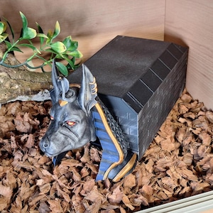 Op de afbeelding: Een zwart en gouden Anubis-hoofdbeeld met blauwe en gouden accenten, rustend op een zwarte bakstenen structuur. Het beeld is omringd door bruine houtsnippers.