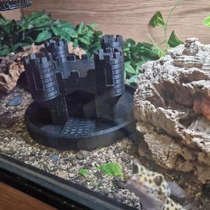 Peut inclure: Une cachette pour reptile en forme de château imprimée en 3D de couleur noire avec une surface texturée. La cachette est placée sur un lit de gravier brun dans un terrarium.