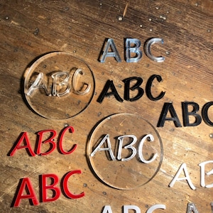 Könnte beinhalten: Eine Sammlung von Acrylbuchstaben, die "ABC" in verschiedenen Stilen und Farben buchstabieren, darunter Schwarz, Weiß, Rot und Klar. Die Buchstaben sind auf einer Holzoberfläche angeordnet.