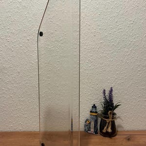 Könnte beinhalten: Transparenter Acrylschildhalter mit abgerundeter Oberseite und drei schwarzen Schraubbefestigungen. Der Halter wird auf einer Holzoberfläche präsentiert. Ein kleiner dekorativer Leuchtturm und ein Glas mit einer Pflanze befinden sich im Hintergrund.