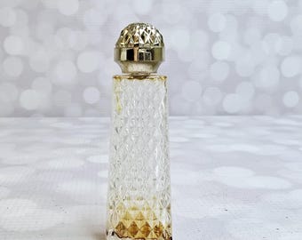Può includere: Un piccolo flacone di profumo in vetro trasparente con un tappo argentato e strutturato. La bottiglia ha un motivo a diamante e contiene una piccola quantità di liquido color ambra. La bottiglia è rettangolare e poggia su una superficie bianca.