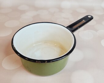 May include: A small, vintage enamel saucepan with a single handle. The pan is olive green with a white interior and a black rim and handle. The handle has a small hole for hanging. A classic kitchenware item.