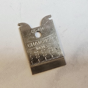 以下が含まれることがあります： 銀色のChampionスパークプラグギャップゲージ。ゲージは長方形で、穴と2つの切り欠き端があります。インチ単位の測定値があり、0.51mmから1.02mmに相当します。